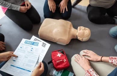 Cpr dummy with a group of students sitting around it while someone is holding a first aid flyer.